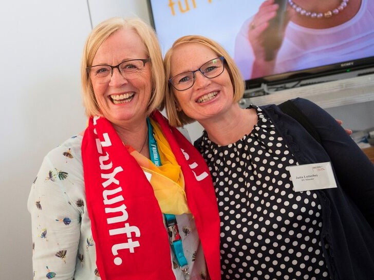 Zwei Frauen der kfd stehen eng beieinander und lachen in die Kamera, eine trägt einen roten Schal mit der Aufschrift „Zukunft“